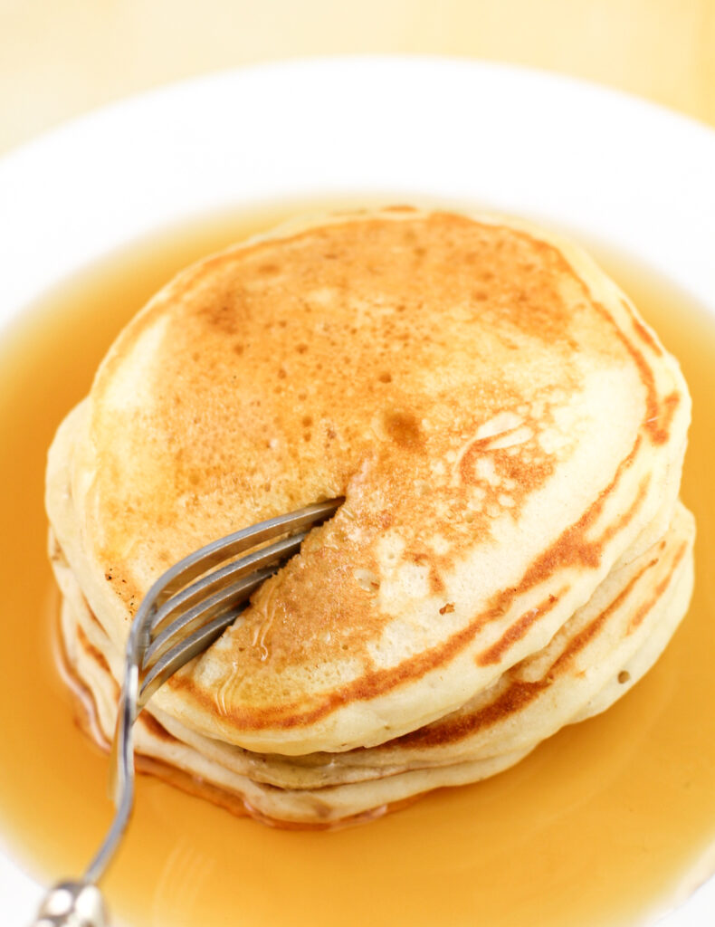 fork pressing into pancakes stacked on white plate with syrup