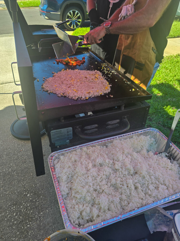 pan of cooked rice in front; large Blackstone griddle in back cooking stir-fry rice