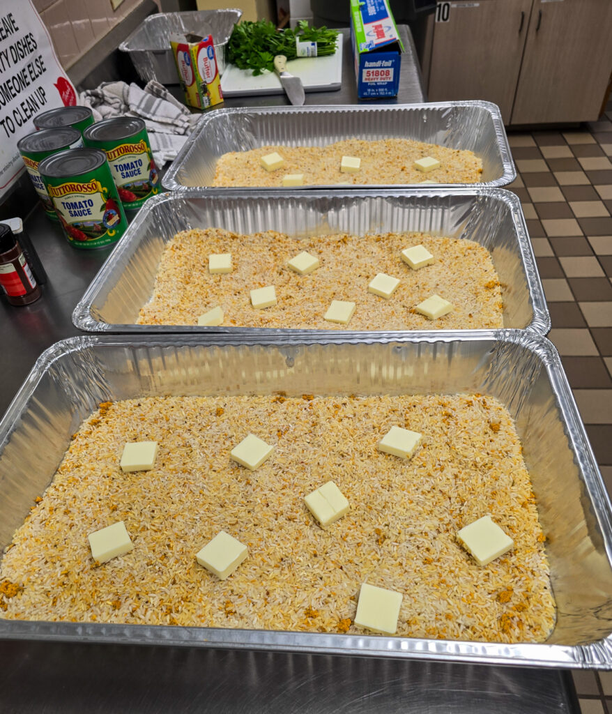 raw Vigo yellow rice topped with pieces of butter in large silver commercial hotel pans for bulk meal preparation.