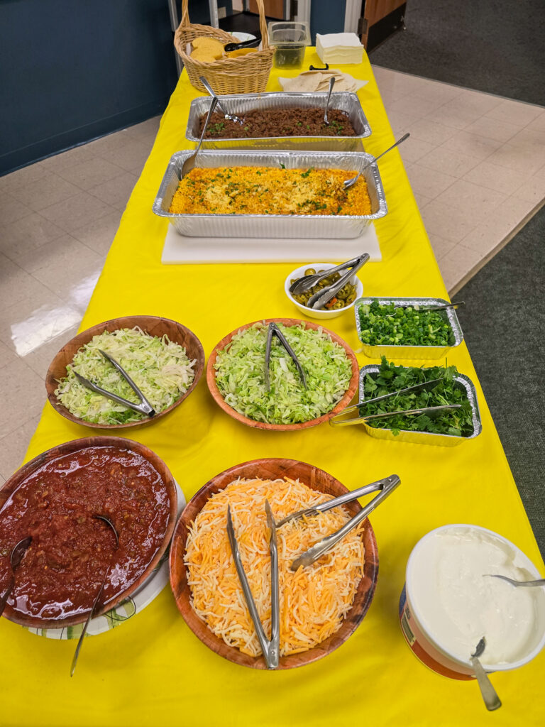 Taco bar buffet set up on a yellow tablecloth with pans of meat, yellow rice, and bowls of toppings, salsa, and tortillas