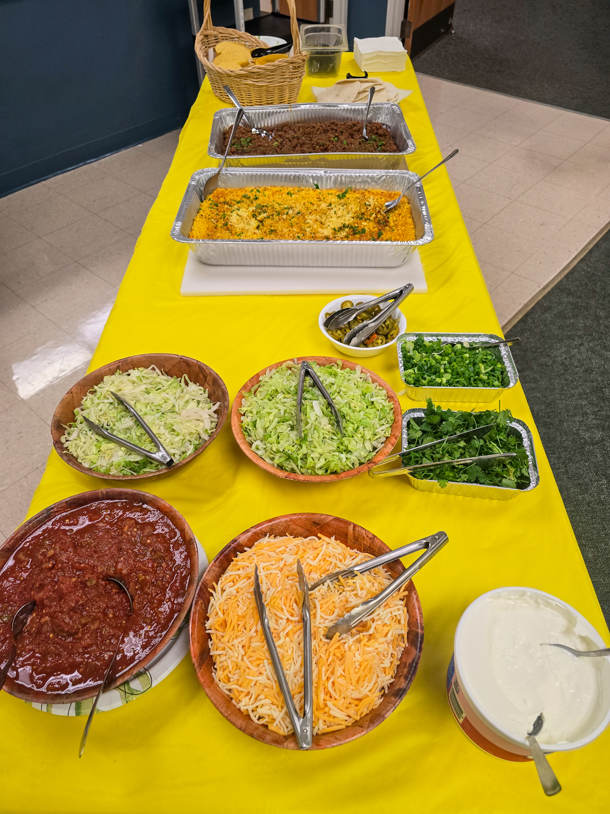 Taco bar buffet set up on a yellow tablecloth with pans of meat, yellow rice, and bowls of toppings, salsa, and tortillas