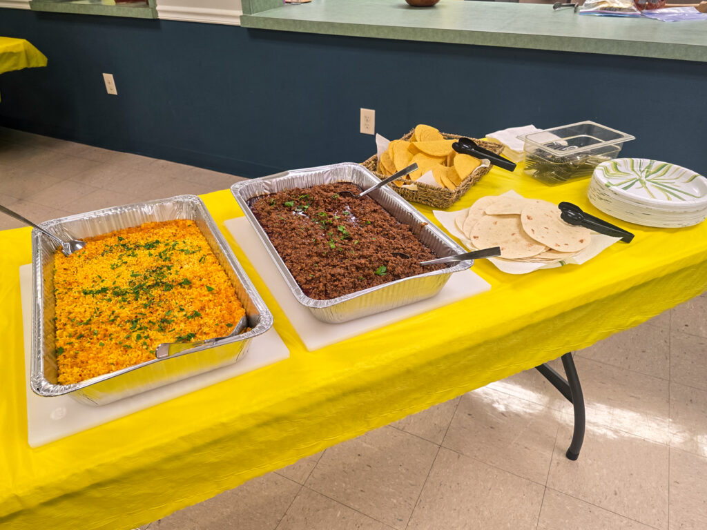hotel pans of yellow rice and taco meat, tortilla shells with tongs, paper plates, and silverware on table with yellow tablecloth