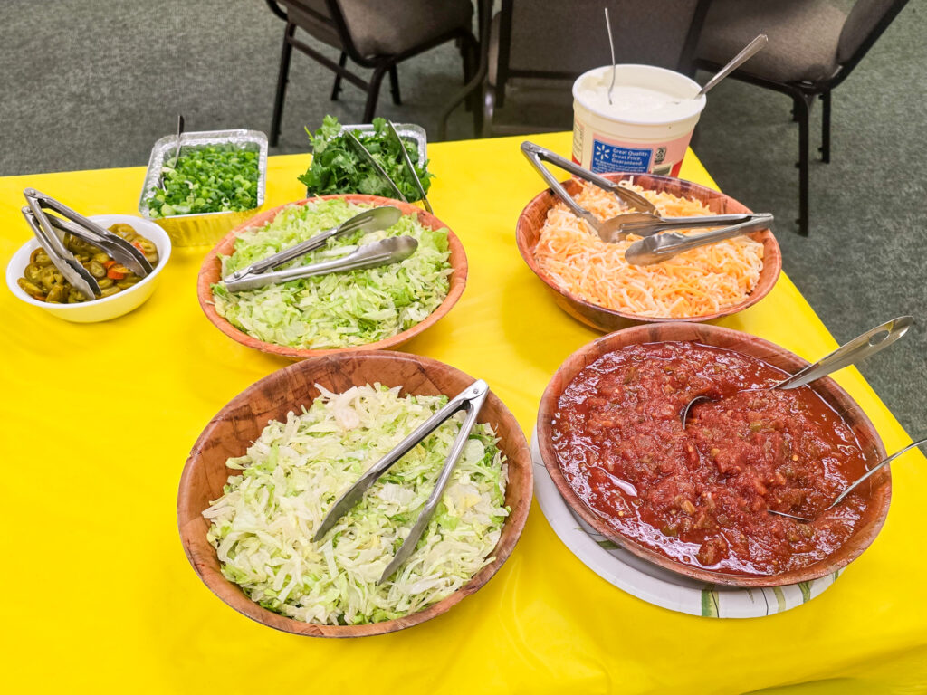 taco fixings (green onion, cilantro, salsa, lettuce, cheese, sour cream, and jalapenos) in bowls on table with yellow table cloth
