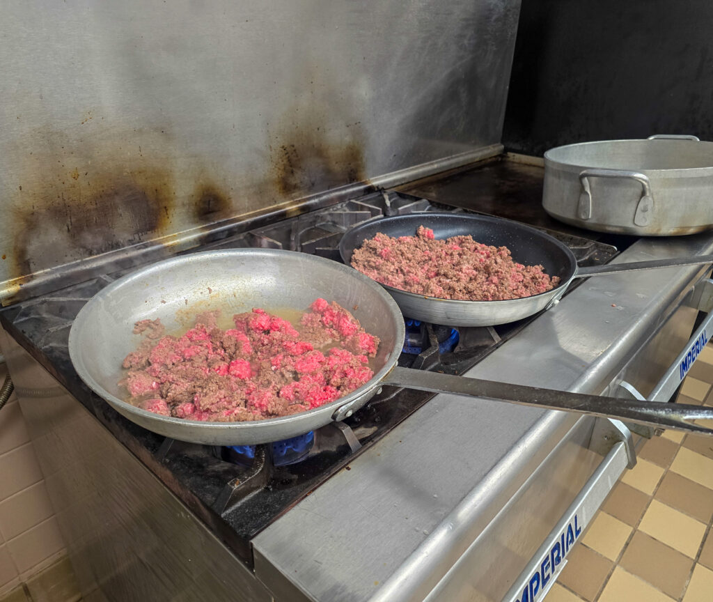 Ground beef browning in two large skillets over gas burners on a commercial stove