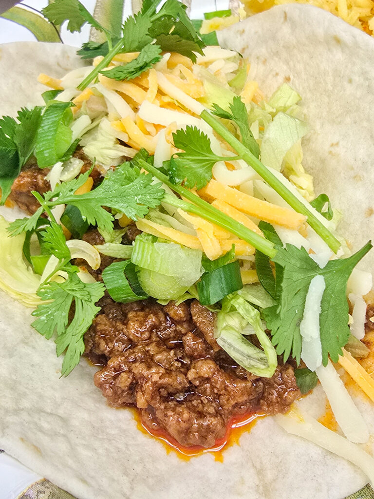 Soft taco filled with seasoned ground beef, shredded cheese, lettuce, green onions, and fresh cilantro