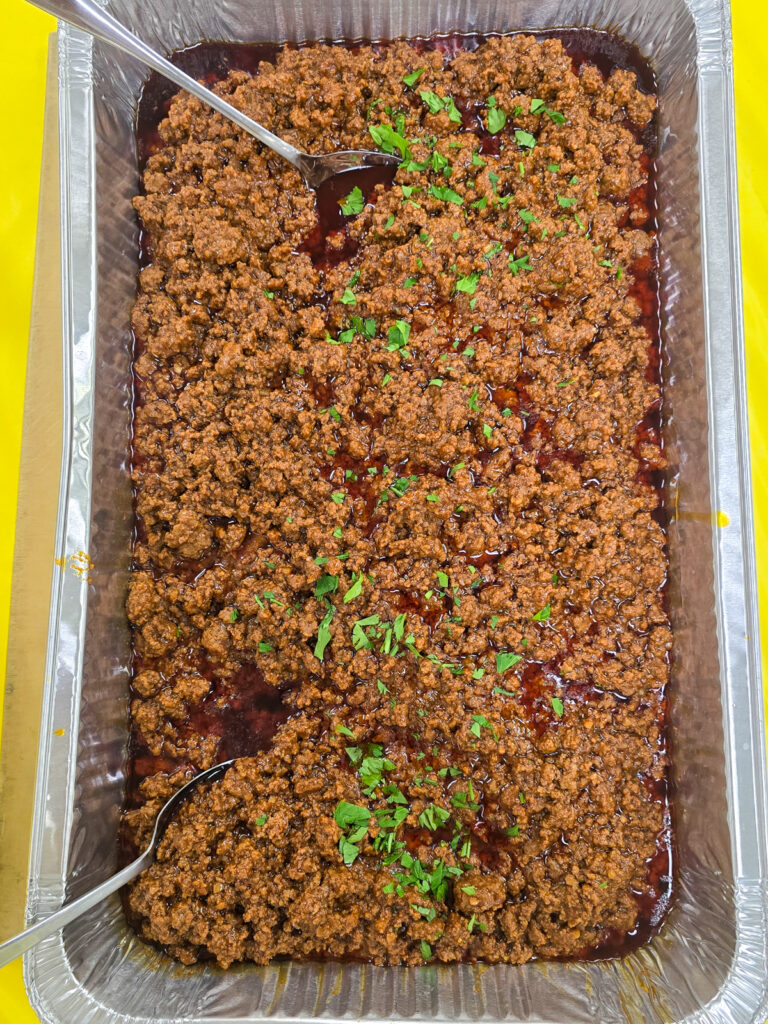 Aluminum catering tray filled with seasoned ground meat garnished with fresh chopped herbs and serving spoons